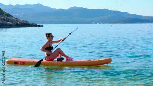 Young woman paddling on a board at sea during summer vacation. Beautiful woman in a bikini enjoying her summer vacation, sitting on a paddleboard and rowing with an oar on the calm turquoise sea