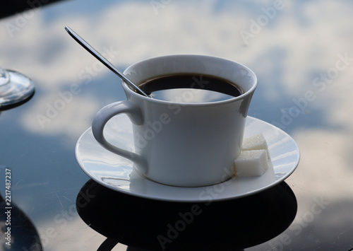 White cup of black coffee with a spoon and sugar cubes on a glossy table, reflecting a bright cloudy sky and creating a calm relaxing atmosphere