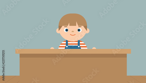 Adorable little boy with a sweet smile playfully peeking over a wooden desk, embodying youthful curiosity and innocent charm in a classroom setting.