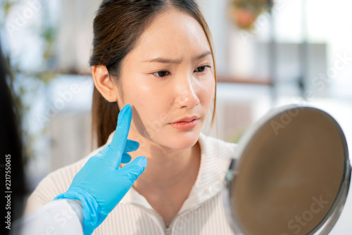 Asian woman patient looking in mirror with anxiety and holding reflection for checking skin problem, aesthetic doctor examining sad face for cosmetic treatment consultation, evaluating skin facial.