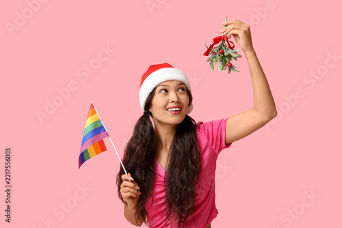 Young Asian woman in Santa hat with LGBT flag and mistletoe on pink backgroun...