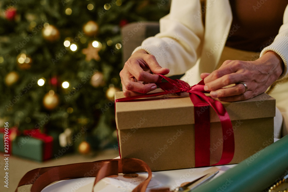 Fototapeta premium Cropped hands of Caucasian adult wrapping holiday present with ribbon near Christmas tree. Suitable for seasonal retail marketing, festive gift giving campaigns, greeting concepts