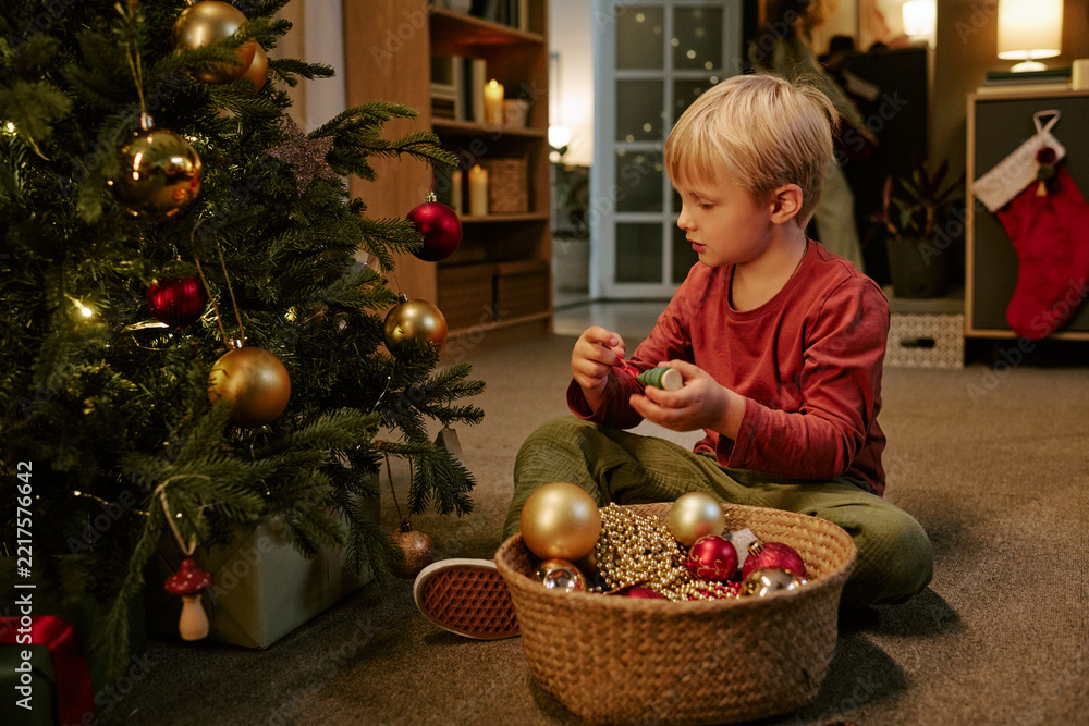 Fototapeta premium Caucasian child decorating Christmas tree with ornaments at home, celebrating winter holidays with family. Useful for seasonal marketing campaigns and holiday promotions