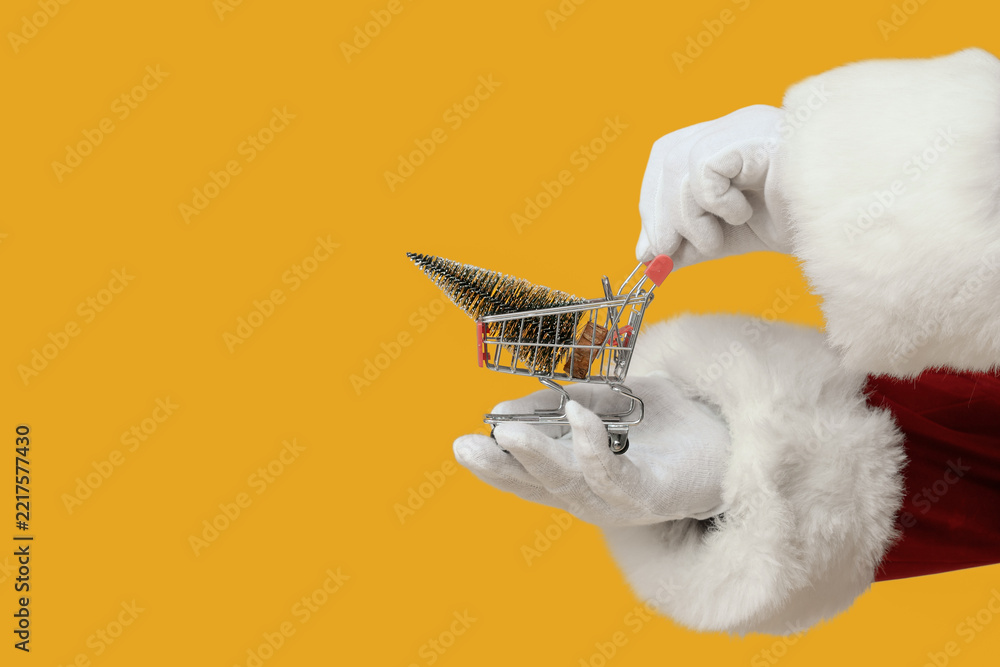 Fototapeta premium Christmas shopping and holiday retail sales, Santa Claus holding a miniature shopping cart with a small pine tree on a solid yellow background