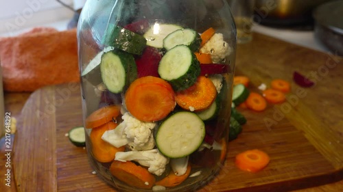 Organic produce preservation with Cucumis sativus in home kitchen. Static close-up shot showing hand adding sliced vegetables into clear glass storage jar.