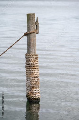 Holzpfahl mit aufgewickeltem Schiffstau steht im ruhigen Wasser eines Hafens bei weichem, bedecktem Tageslicht in maritimer, stilvoller Atmosphäre.