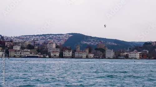 Scenic view of Istanbul and the Bosphorus Strait from a moving boat, with waterfront buildings, seagulls and a dramatic cloudy sky in the background.