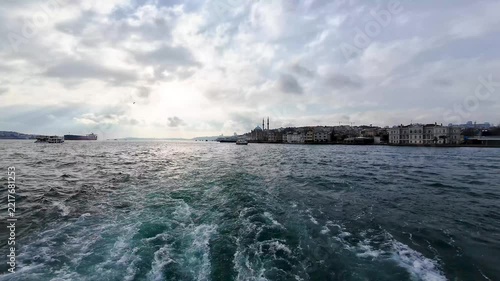 Scenic view of Istanbul and the Bosphorus Strait from a moving boat, with waterfront buildings, ferries and a dramatic cloudy sky in the background.
