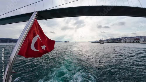 Turkish flag waving from a boat cruising along the Bosphorus in Istanbul, with the Bosphorus Bridge, waterfront and city skyline in the background.