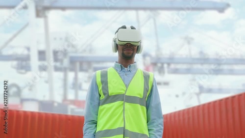 Smiling African American male wearing virtual reality headset and reflective vest while standing near shipping containers. Interacting with digital interface. Zooming in on image in VR technology.