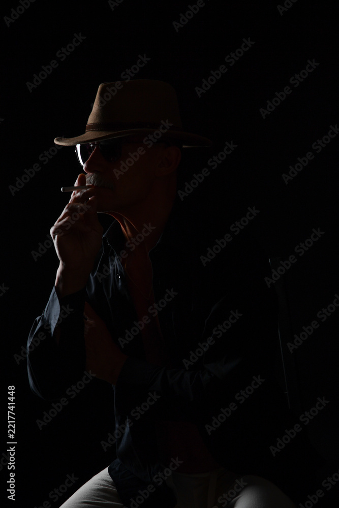 Naklejka premium Dramatisches Low-Key-Porträt eines älteren Mannes mit Hut, Zigarette und dunkler Kleidung vor schwarzem Hintergrund. Vintage-Stil, französisches Flair, Lebensstil und Charakterfotografie.