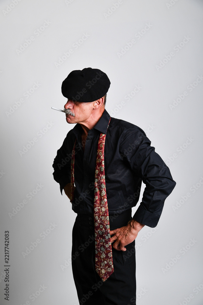 Naklejka premium Charakterporträt eines älteren Mannes mit schwarzer Schiebermütze, schwarzem Hemd, gemusterter Krawatte und Zigarette in selbstbewusster Pose. Vintage-Stil, französischer Charme und Studiofotografie.