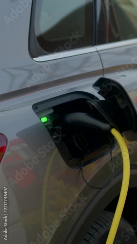Close-up of electric car side charging inlet showing yellow cable plugged in, and green status light glowing on grey bodywork.