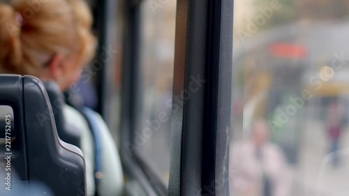 Blonde woman with bun seated on city bus, gazing at blurred street through window as daylight enters quiet cabin.