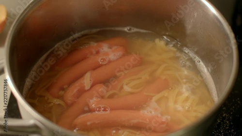 Close-up view of sausages and spaghetti boiling together in stainless steel pot on stovetop. Warm kitchen light highlights pale orange sausages partially submerged in cloudy water.
