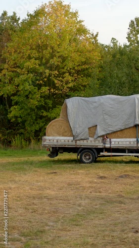 Parked flatbed truck sits on grassy field, carrying large round hay bales covered with weathered tarp, with nearby trees starting autumn colors.