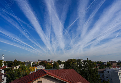 Dramatic pattern of crisscrossing airplane contrails spreading across a clear blue sky over suburban rooftops during bright daylight in an atmospheric cityscape