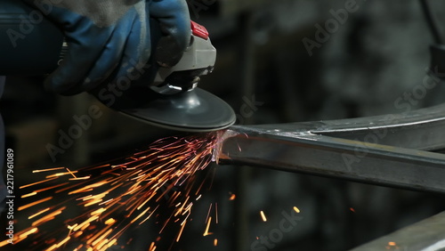 Worker uses angle grinder to shape steel component in plant workshop closeup. Hot sparks scatter across frame. Metalworking industry concept