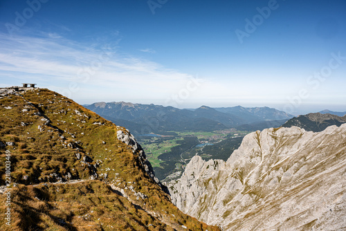 High angle panoramic view from the rocky peaks of the Karwendel Alps featuring a wooden bench on a grassy ridge overlooking the valley below.
