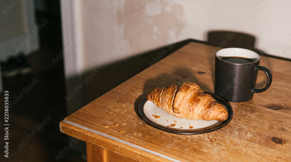 Naklejka premium Fresh croissant on a plate with a cup of coffee on a rustic wooden table, cozy breakfast scene with warm light and copy space