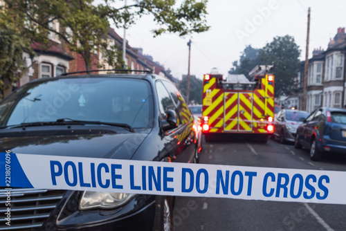 Police tape blocking a residential street with emergency vehicle and fire engine in the distance