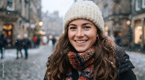 Young woman smiling in a city street during a winter snowfall, wearing a white beanie and a chunky knit scarf, with blurred urban background