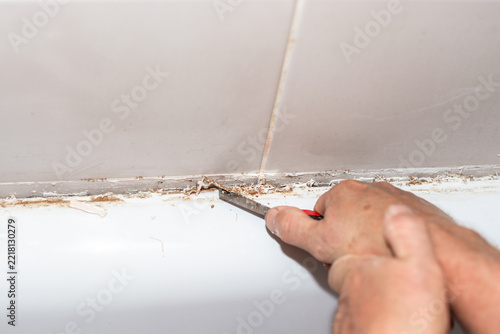 A worker uses a sharp scraper tool to remove old dirty silicone sealant from a bathtub edge. DIY residential bathroom restoration process. Home improvement theme.