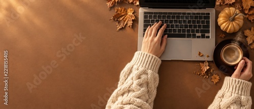 Hands in cozy cable knit sleeves using a laptop on a brown desk with pumpkins, autumn leaves, and steaming coffee for home office and blog mockup