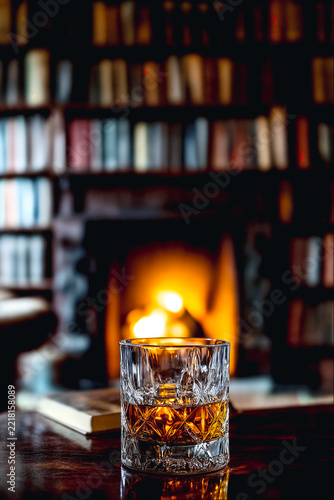 Glass of whiskey on a table in front of a cozy fireplace and bookshelves in a dim library room