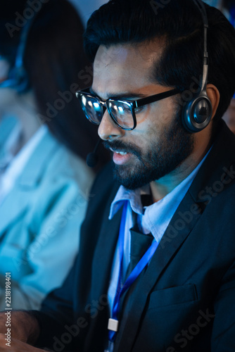Fotografie Indian support agent talks with a customer through a headset while working late hours at an office desk to resolve technical issues and provide online assistance to clients