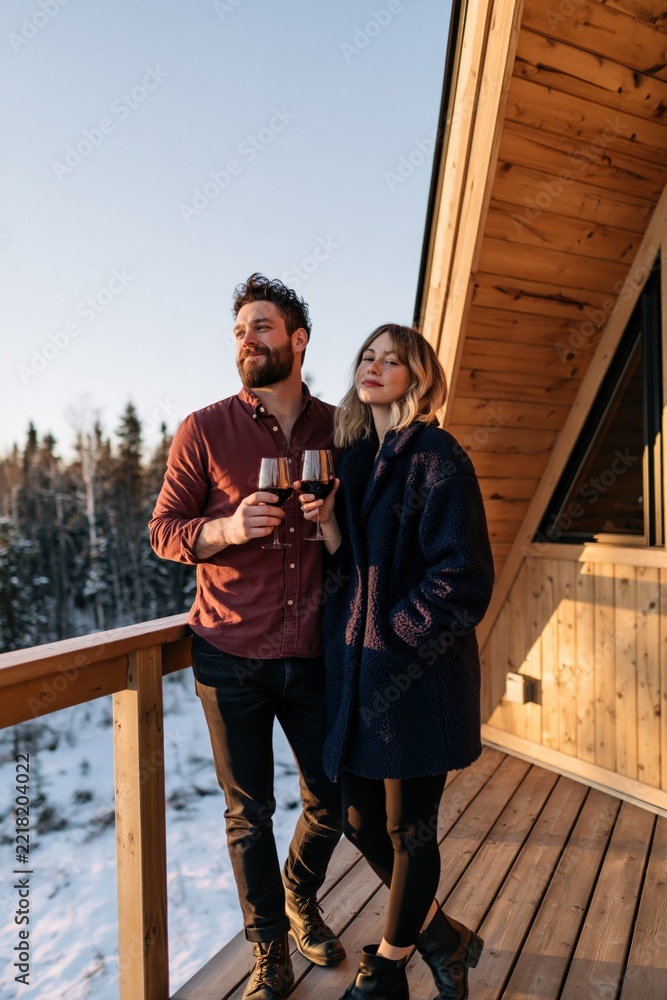 Fototapeta premium Couple on wooden cabin balcony holding wine glasses overlooking snowy forest for romantic retreat background and cozy lifestyle banner