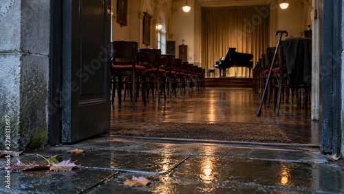 An open doorway reveals a room with rows of wooden chairs, a black grand piano in the background, and a walking cane leaning against a chair