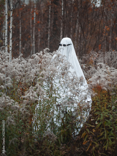 A ghost in a white sheet and sunglasses is walking in the autumn forest.