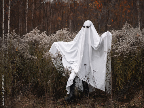 cheerful ghost in sunglasses and a white sheet is posing in an autumn forest.