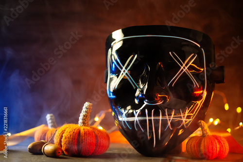Black glowing mask on a table with knitted autumn pumpkins, smoky background