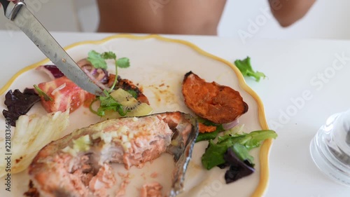 Hands cutting kiwi and salad with serrated knife beside partially eaten salmon and roasted sweet potato on white plate with yellow rim, low angled close-up at home dining table