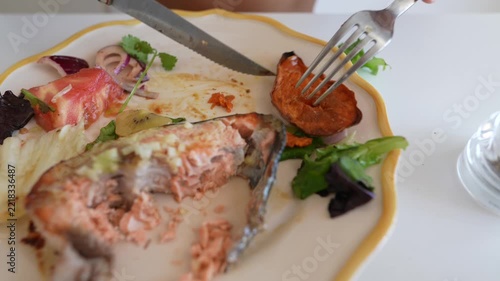 Close-up of fork and serrated knife cutting and lifting roasted sweet potato and salmon from scalloped white plate at dining table with tomato wedge and salad leaves
