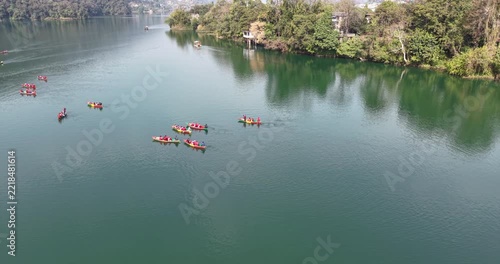 Turning aerial view reveals several small, vibrant red boats filled with people gliding across a calm lake, surrounded by lush, forested hills.