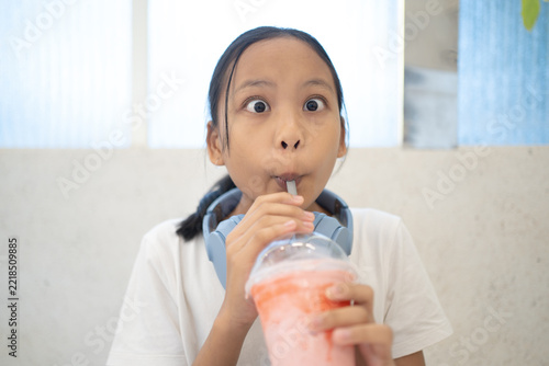 Funny Asian girl wearing headphones making a surprised face with wide eyes while drinking a sweet pink iced smoothie