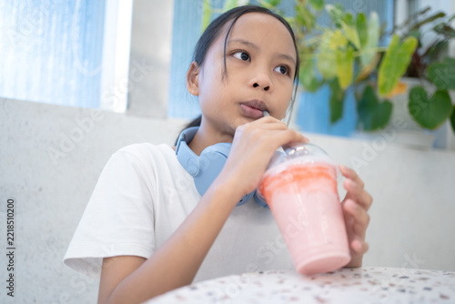 Relaxed Asian tween girl wearing blue headphones around her neck drinking a sweet pink iced smoothie from a plastic cup in a modern cafe