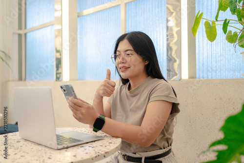 Happy Asian woman with glasses showing thumbs up gesture while using a smartphone and laptop in a cafe