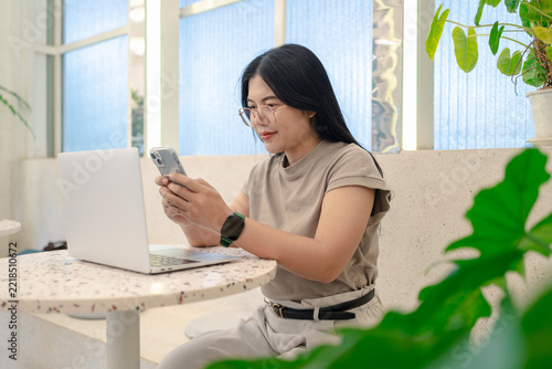 Smiling Asian freelance woman with glasses happily using a smartphone sitting with a laptop in a modern cafe
