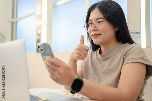 Smiling Asian woman with glasses showing thumbs up gesture while using smartphone and laptop in a cafe