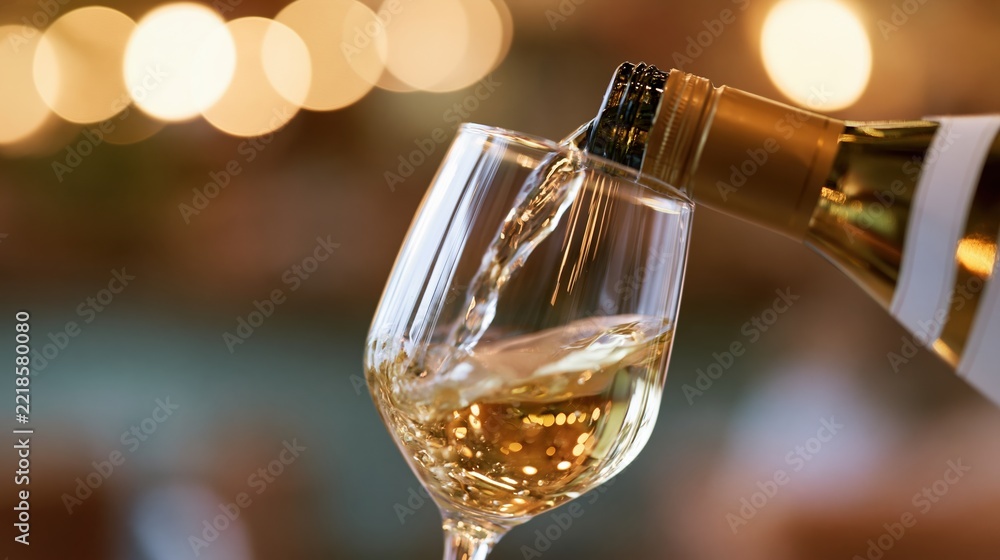 Fototapeta premium White wine being poured from a gold-labeled bottle into a clear glass at a dimly lit indoor setting