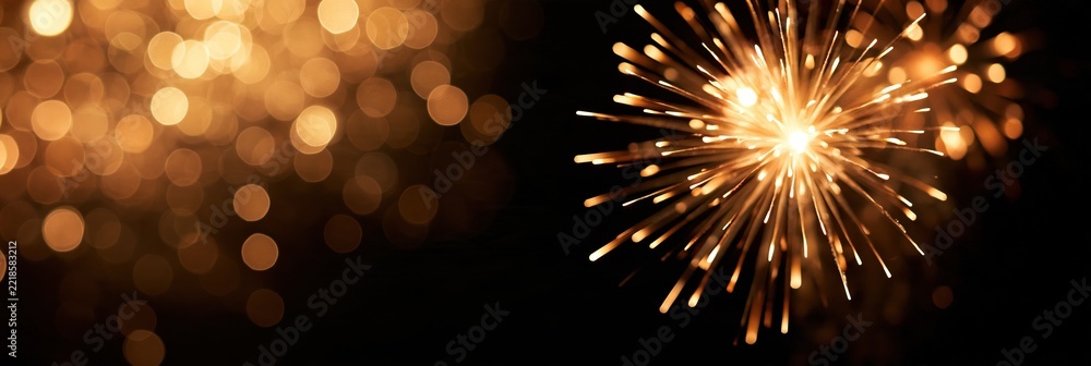 Fototapeta premium Golden sparkler bursts against black background with warm bokeh lights and generous copy space, creating elegant festive celebration banner for New Year, holiday party, anniversary and joyful events