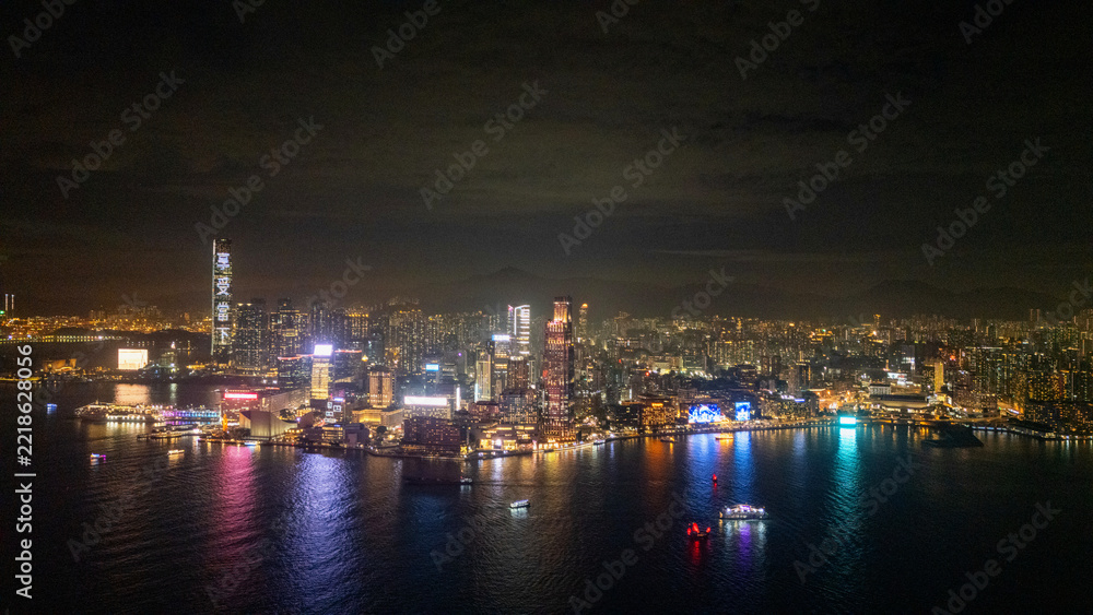 Fototapeta premium Aerial drone view of the Tsim Sha Tsui skyline in Hong Kong at night, neon waterfront buildings glowing across the black water of Victoria Harbour with boats passing