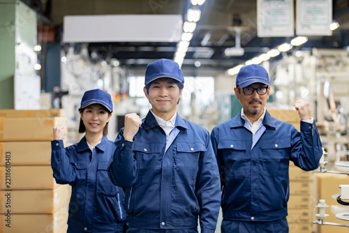 製造工場で笑顔でガッツポーズをする若手男性と女性、ベテラン男性の3人の作業員チーム