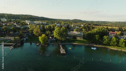 A drone pulls back and rises over Lake Starnberg in Germany, widening the view of the tree-lined waterfront, piers, boats and lakeside buildings.