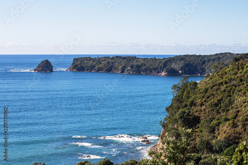 A rugged forested headland rises above the deep blue ocean in Coromandel, with a small rocky island and clear summer sky completing the serene coastal scene