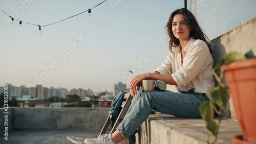 Young woman sitting on urban rooftop at sunset, peaceful female relaxing with coffee cup on city terrace, casual lifestyle portrait